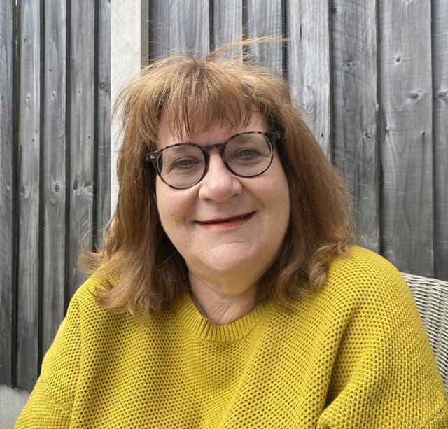 a woman with brown hair and glasses, wearing a mustard coloured jumper