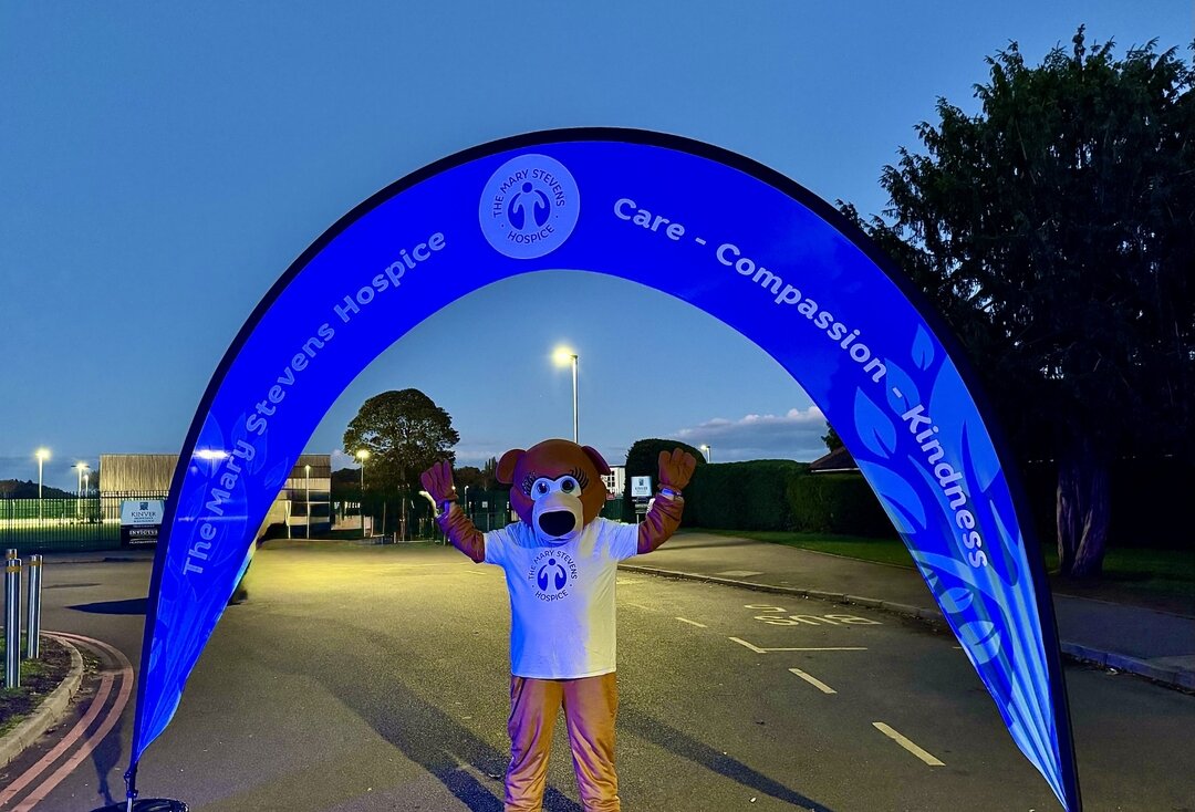 A mascot dressed as a bear, wearing a branded t-shirt, stands beneath a large, glowing blue arched banner for The Mary Stevens Hospice