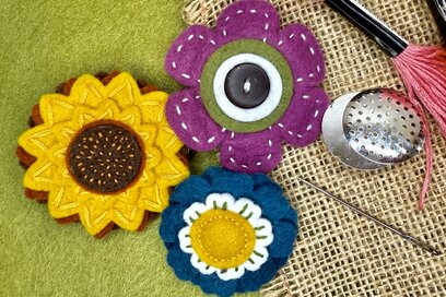 three colorful handmade felt flower brooches stitched with decorative details on a burlap and green felt background, alongside sewing tools including a needle and thread