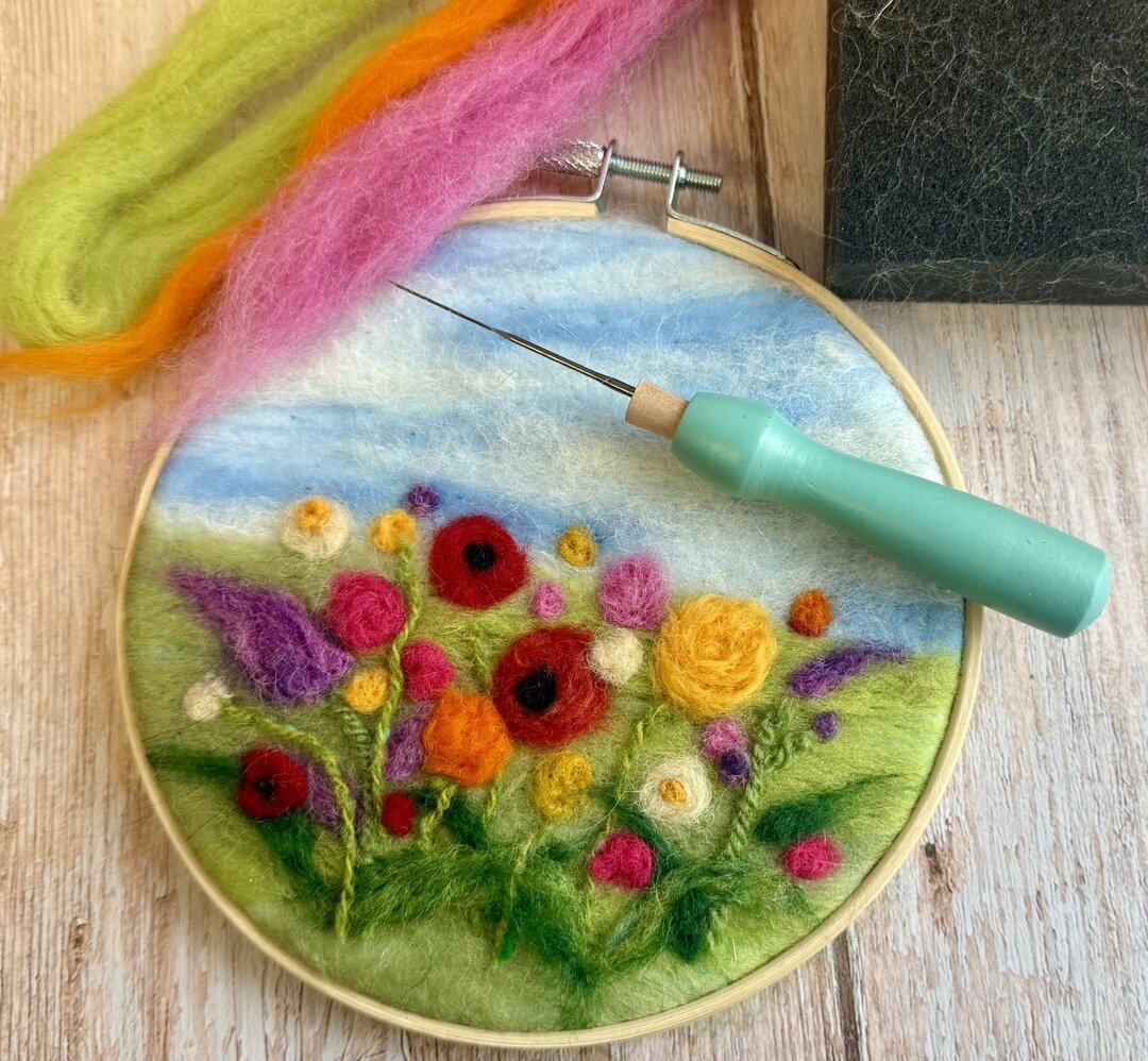 Needle felting project in an embroidery hoop showing colourful flowers, with felting needle tool and wool fibres on a wooden surface.
