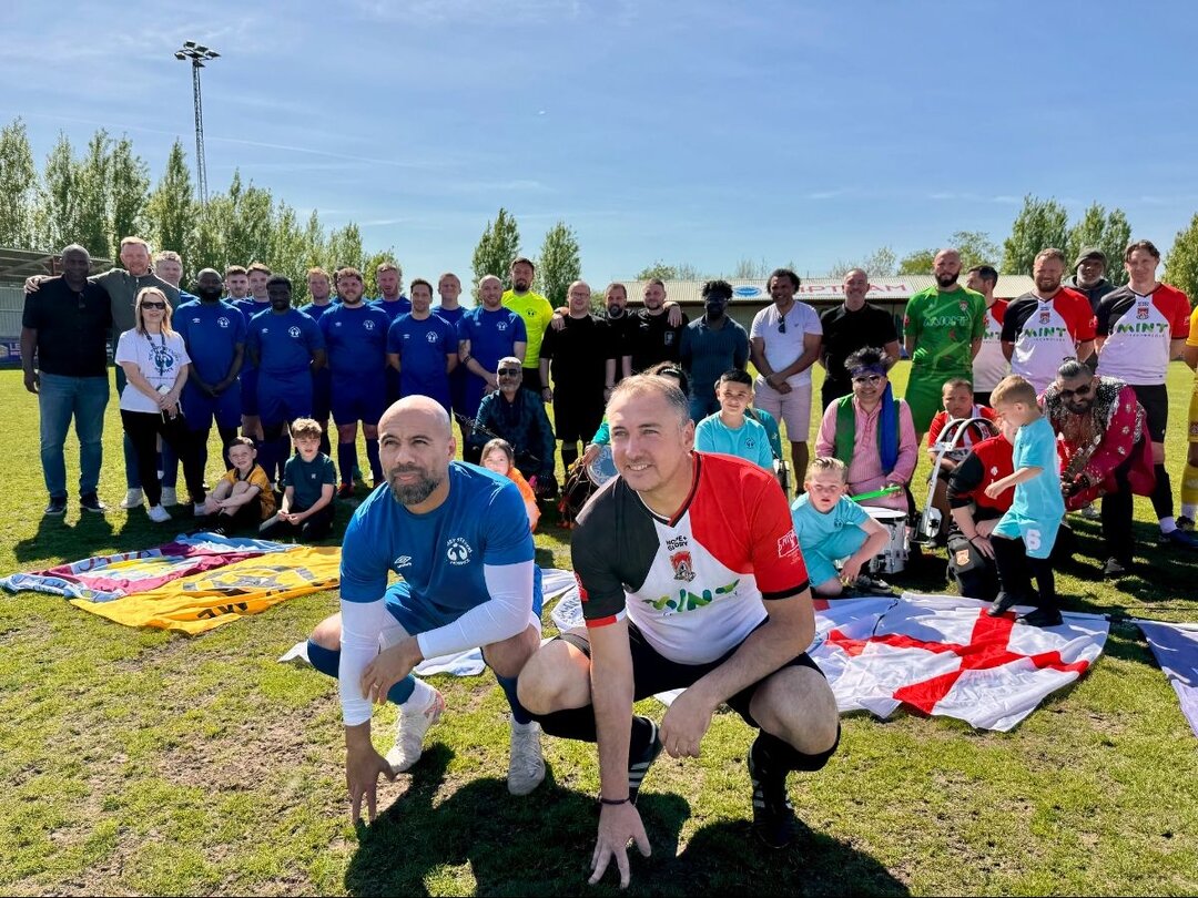 Group of football players, families, and children posing together on a sunny pitch during a charity match.