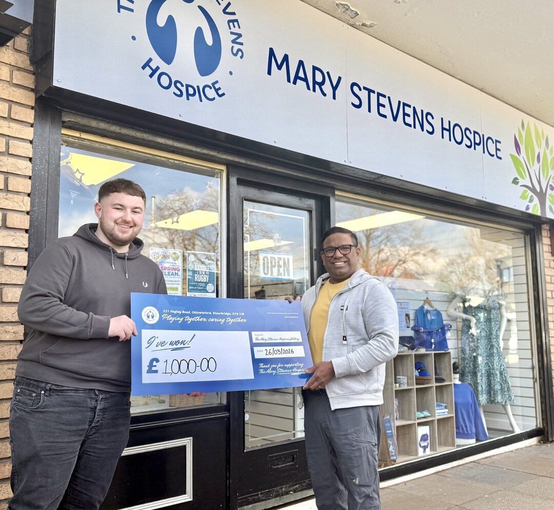 two men infront of a mary stevens hospice charity shop, holding a large cheque for £1,000