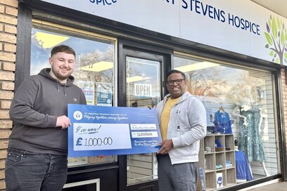 two men standing in front of a mary stevens hospice charity shop, holding a large cheque for £1,000