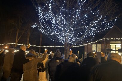 A group of people holding candles gathered outdoors at night beneath trees adorned with glowing fairy lights
