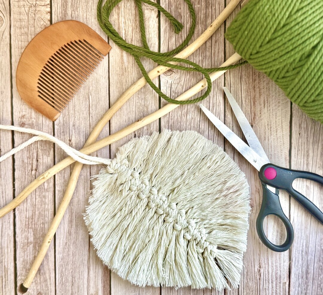 Flat lay of macramé craft materials including green yarn, wooden comb, sticks, scissors, and a partially finished woven piece on a wooden surface.