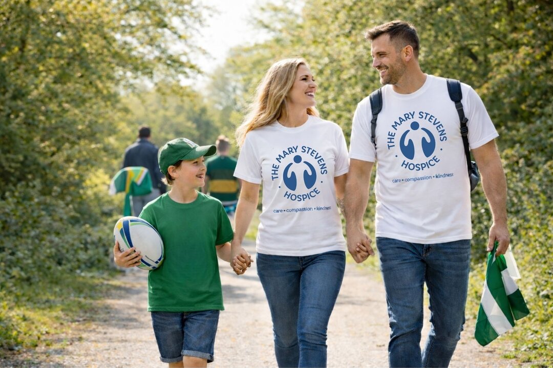 happy, smiling family walking, with the mary stevens hospice t-shirts on. a young boy is holding a rugby ball in a green t-shirt. they are walking through a park area
