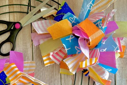 Colourful fabric strips tied onto a wire wreath frame, with scissors nearby on a wooden surface, creating a bright handmade decoration.