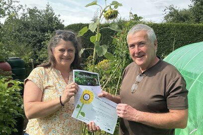 two people standing in an allotment with a tall sunflower, holding a sunflower growing competition certificate
