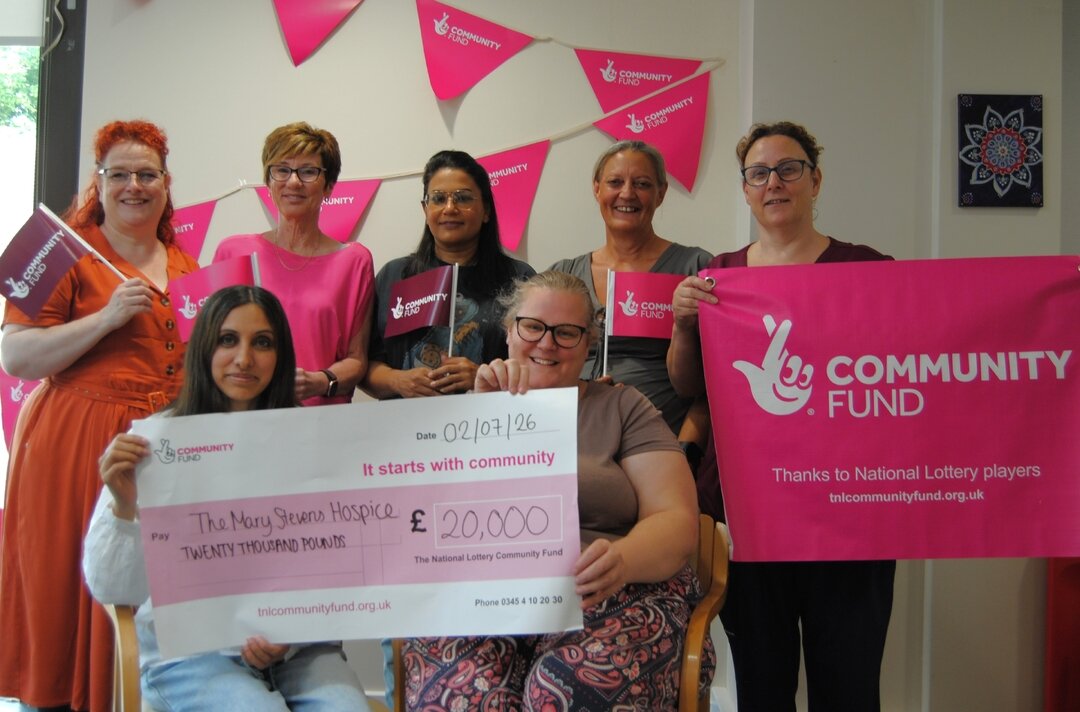 Staff and patients from The Mary Stevens Hospice smiling and holding National Lottery flags, with two people seated at the front holding a large cheque for £20,000 from The National Lottery Community Fund, against a backdrop of pink bunting and a Community Fund banner