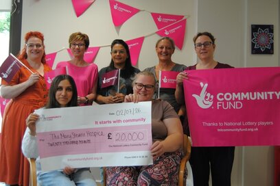 Staff and patients from The Mary Stevens Hospice smiling and holding National Lottery flags, with two people seated at the front holding a large cheque for £20,000 from The National Lottery Community Fund, against a backdrop of pink bunting and a Community Fund banner