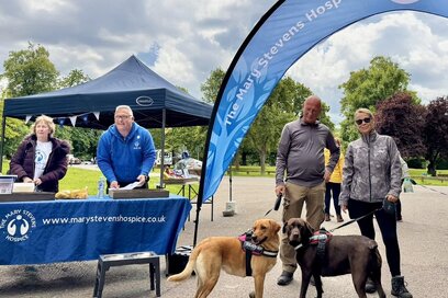At an outdoor event for The Mary Stevens Hospice, a man and woman stand with two large dogs on leashes next to a blue information booth. The booth is manned by two volunteers, and a large blue feather flag stands nearby against a backdrop of green trees and a cloudy sky.