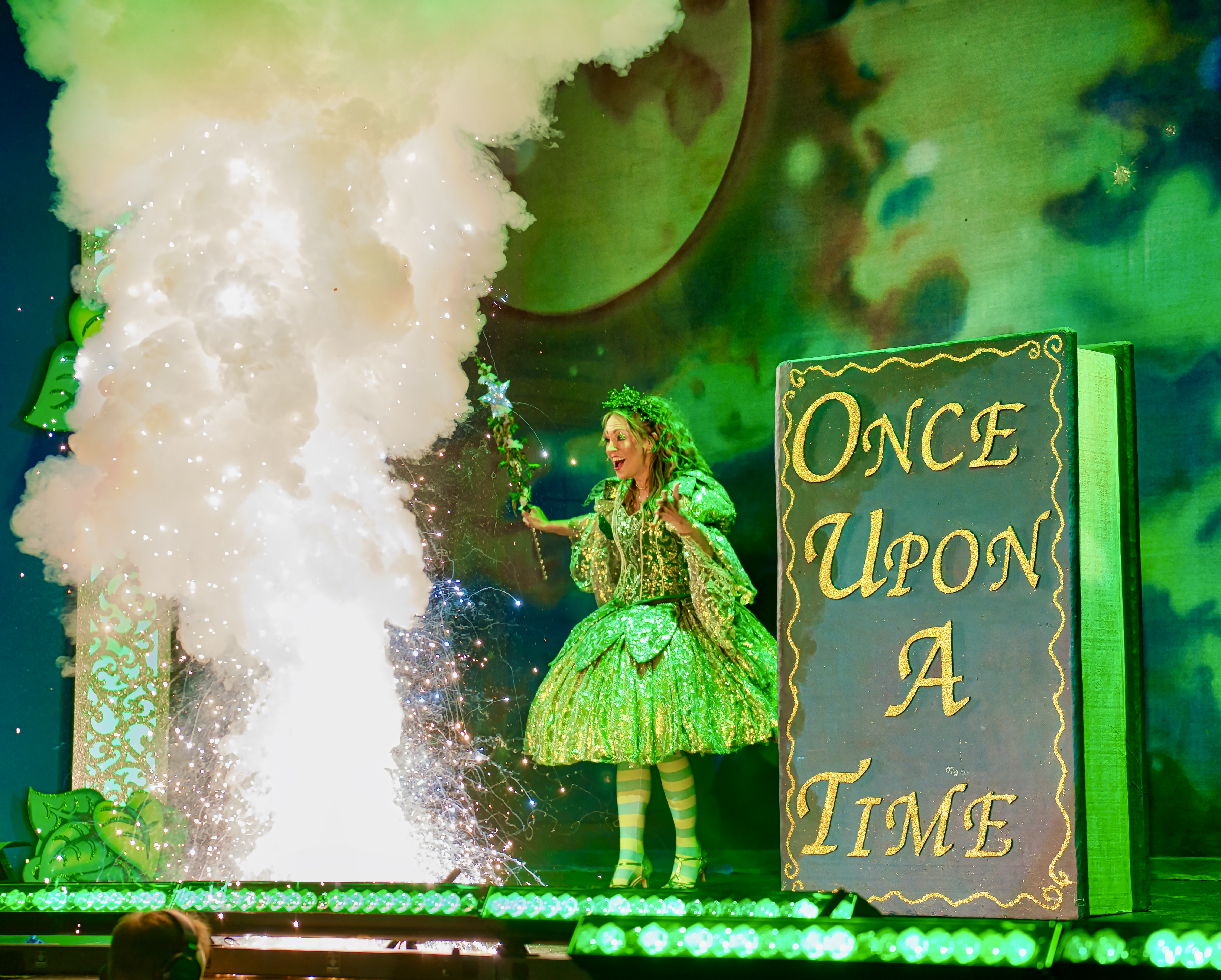 Green-lit pantomime scene with a fairy in a sparkling emerald dress conjuring smoke and sparks on stage beside a giant book reading &ldquo;Once Upon a Time.&rdquo;