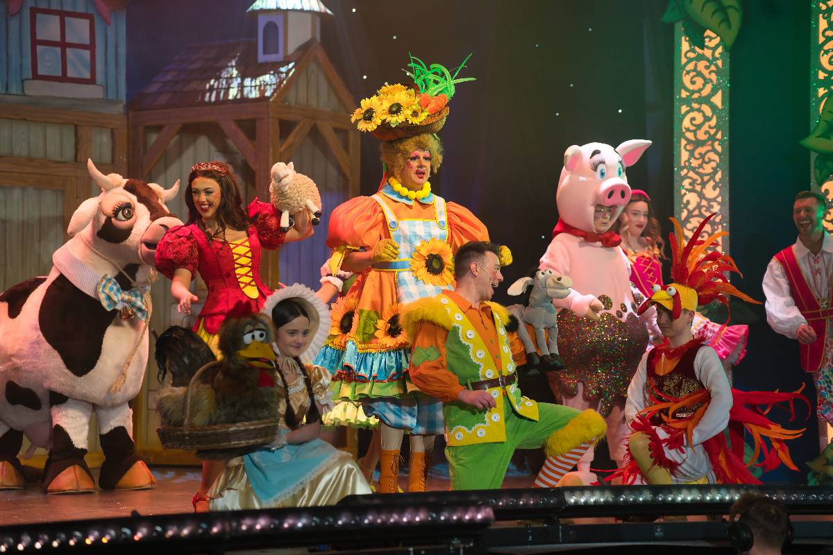 Colourful Jack and the Beanstalk pantomime scene with costumed villagers, animals, and fairy-tale characters gathered together on a lively village stage.